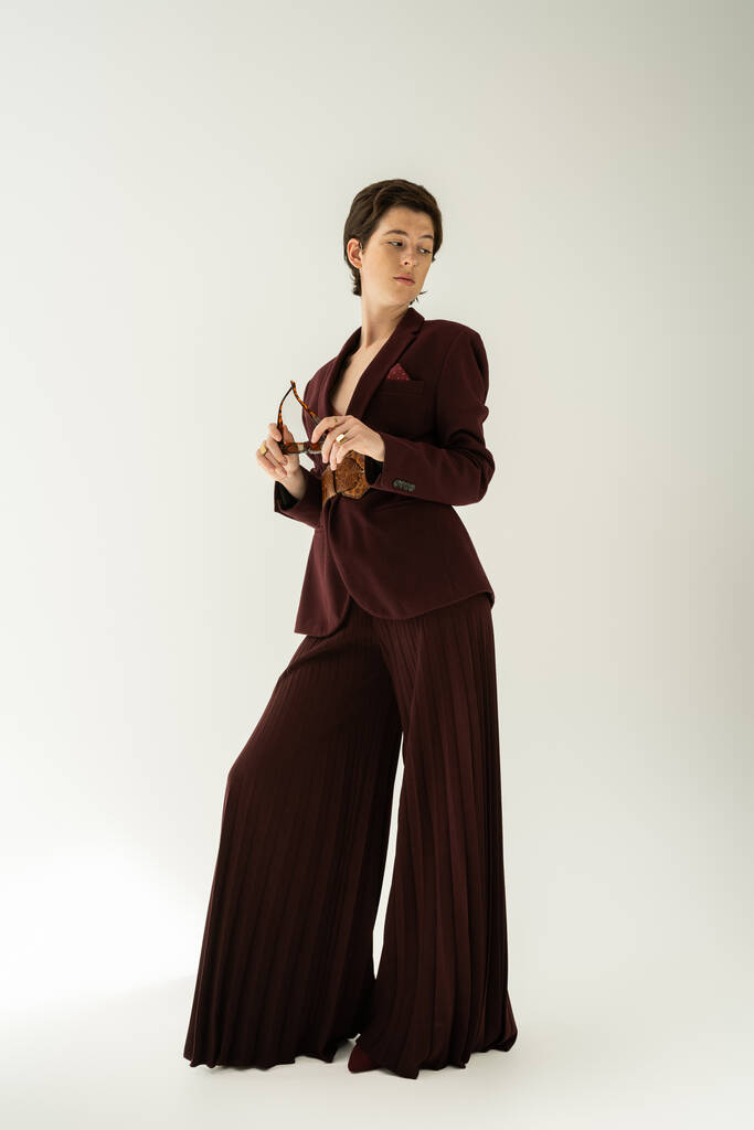 full length of brunette woman in elegant pantsuit holding sunglasses and looking away on grey background - Photo, Image