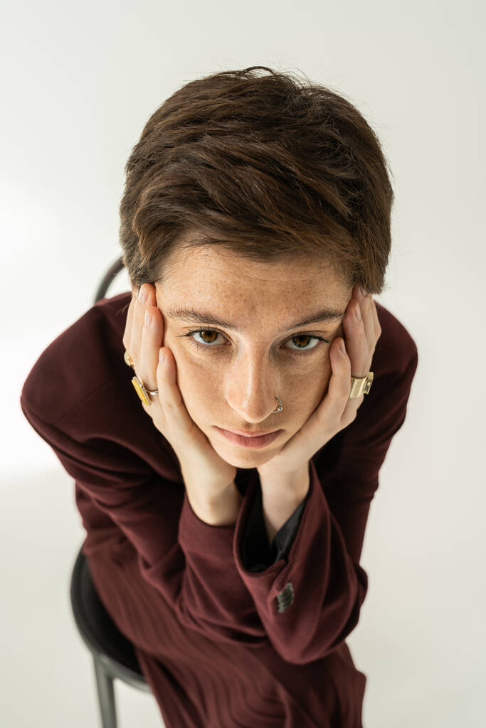 vista de ángulo alto de la mujer pecosa reflexiva en blazer y anillos de dedos tomados de la mano cerca de la cara y mirando a la cámara en gris  - Foto, imagen