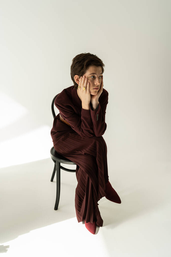 Geniş pantolonlu, modaya uygun ve düşünceli bir kadın sandalyede oturuyor ve gri arka plana bakıyor. - Fotoğraf, Görsel