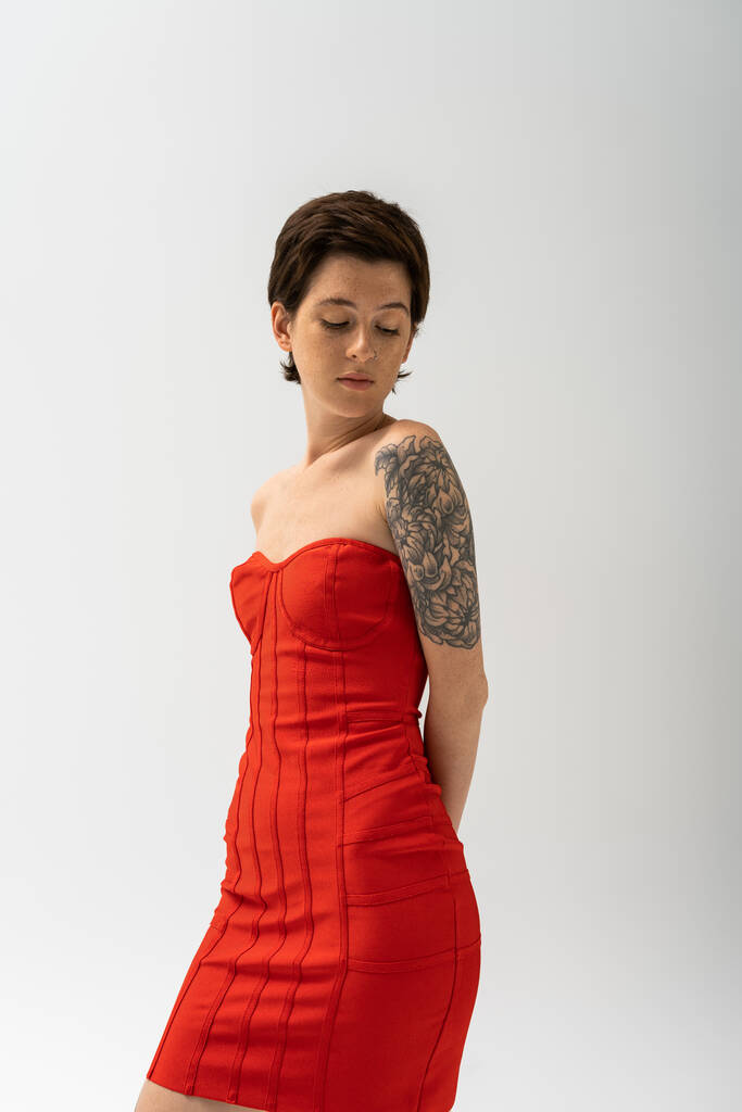 mujer tatuada de moda en vestido rojo sin tirantes posando con las manos detrás de la espalda aisladas en gris - Foto, imagen