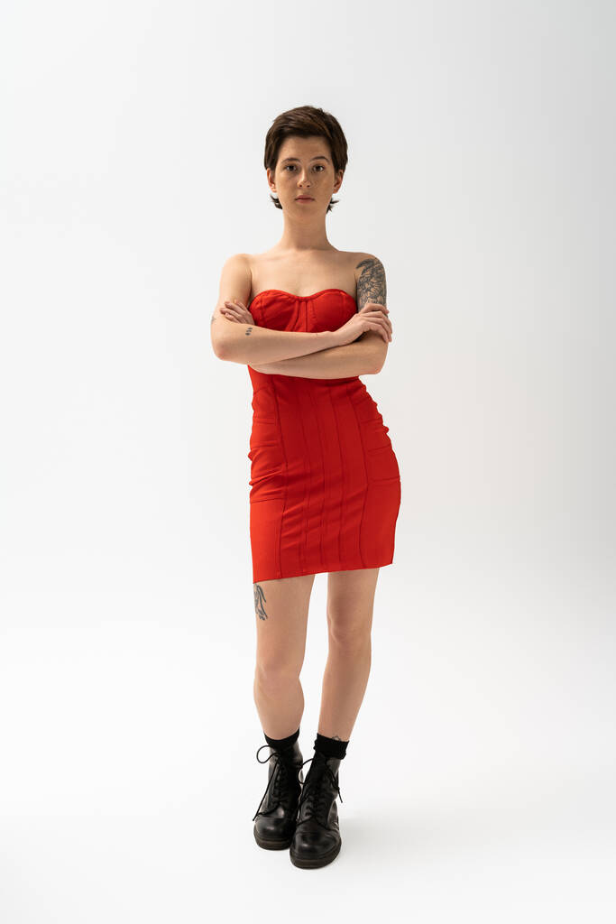 スレンダーな入れ墨の女性の完全な長さで赤ストラップレスドレスと黒ブーツポーズでクロスドアームズオングレー  - 写真・画像