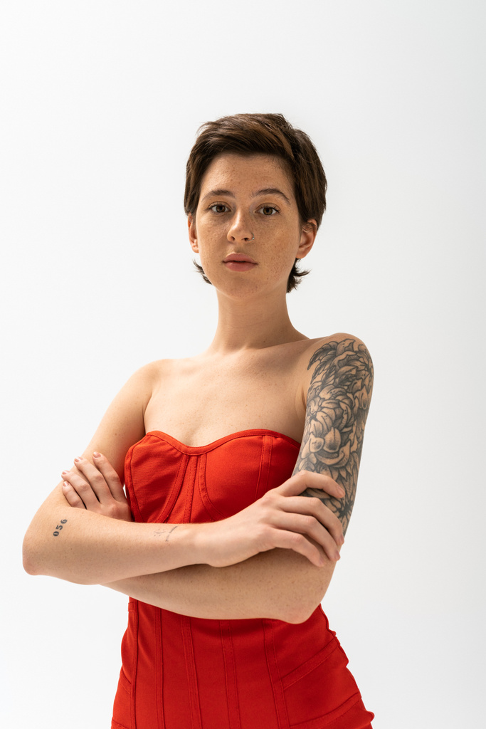 νεαρή γυναίκα με τατουάζ σε κόκκινο στράπλες φόρεμα σταυρώνουν τα χέρια και κοιτάζοντας κάμερα σε γκρι φόντο - Φωτογραφία, εικόνα