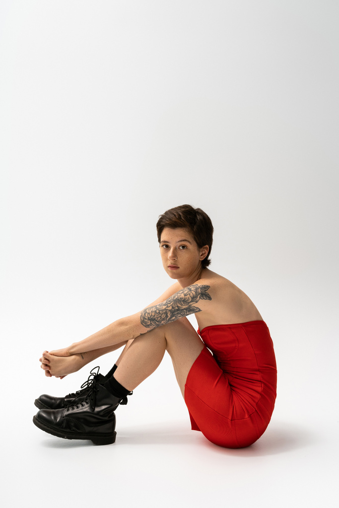 full length of brunette tattooed woman sitting in red corset dress and rough boots on grey background - Photo, Image