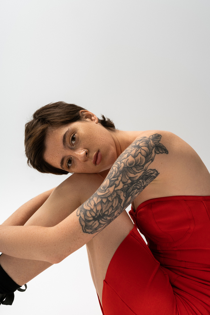 мечтательная татуированная женщина в красном корсетном платье сидит и смотрит в камеру на сером фоне - Фото, изображение