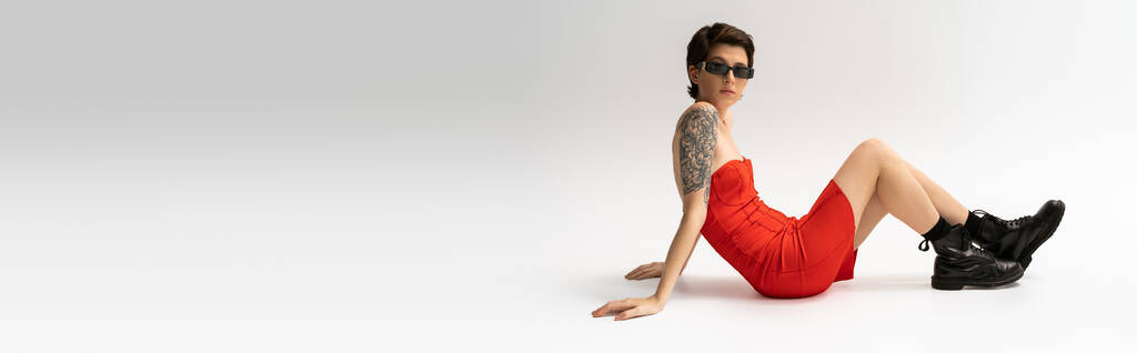 longitud completa de mujer delgada tatuada en botas negras y vestido de corsé rojo sentado sobre fondo gris, pancarta - Foto, Imagen