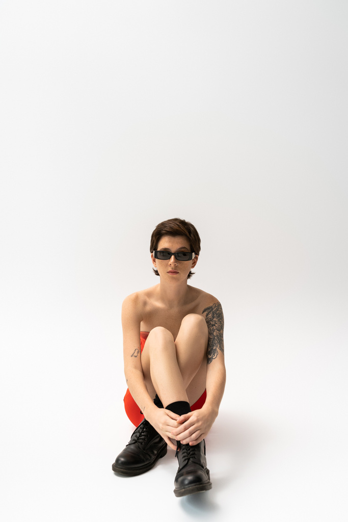 longitud completa de mujer joven tatuada en botas negras ásperas y gafas de sol de moda sentadas sobre fondo gris - Foto, Imagen