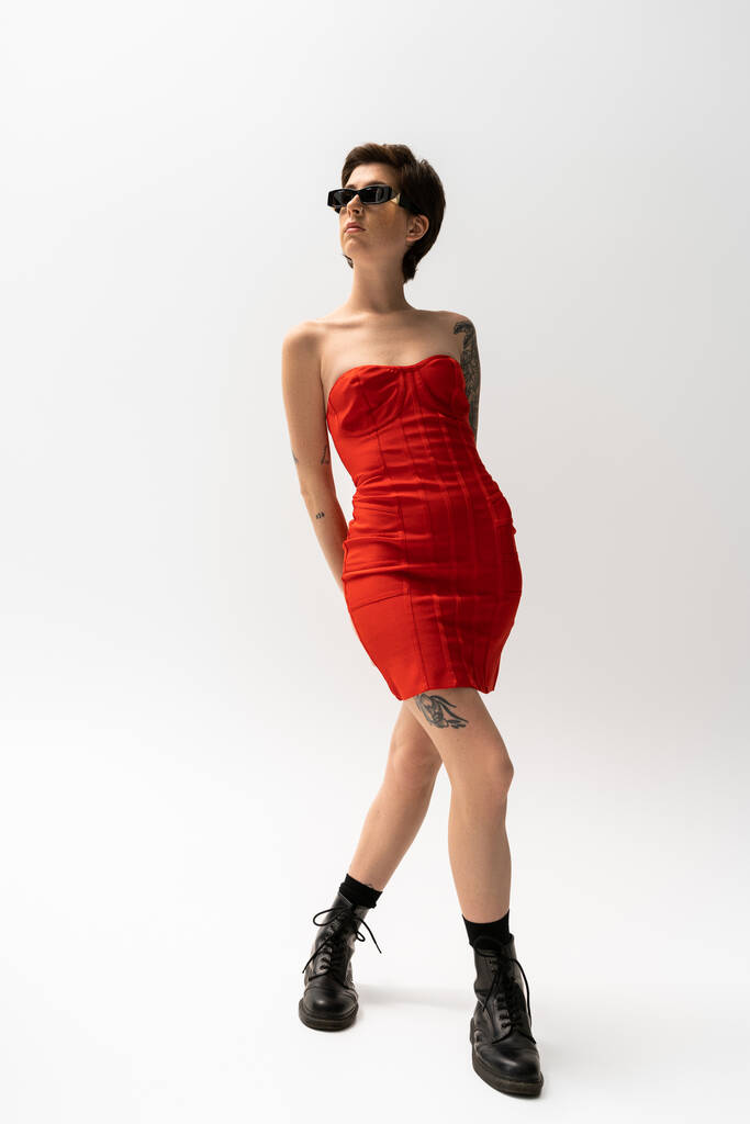 teljes hossza tetovált nő elegáns fűző ruha és napszemüveg pózol szürke háttér - Fotó, kép