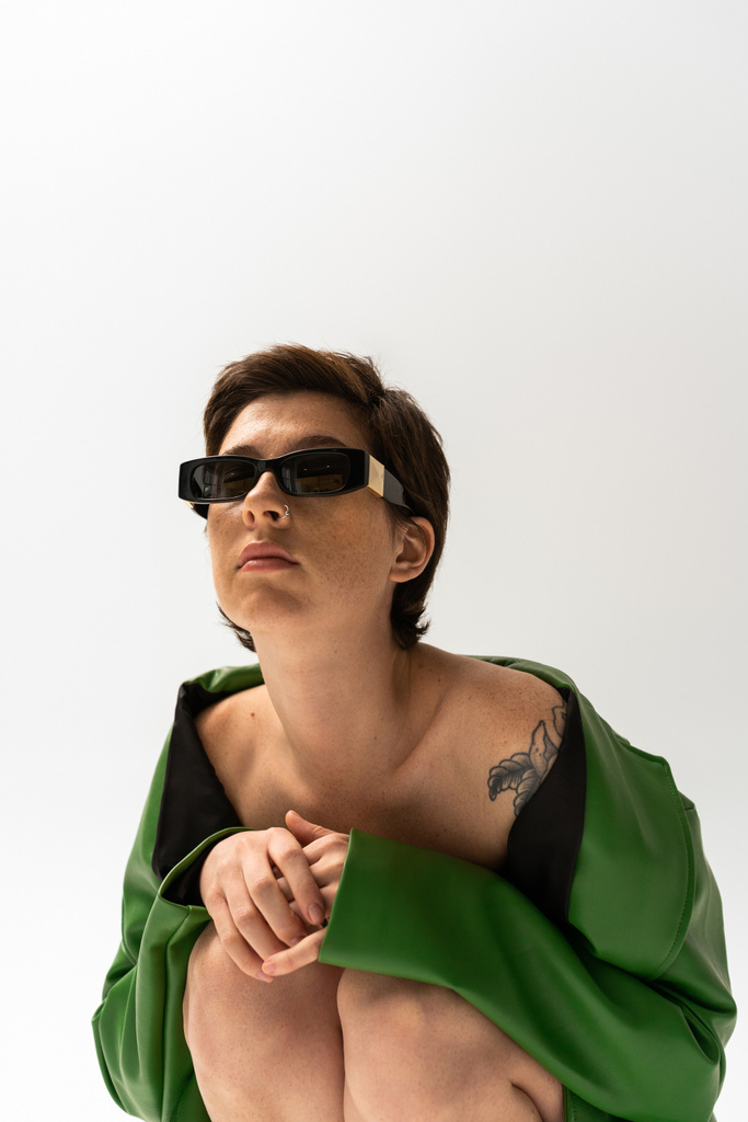 wytatuowana kobieta w zielonej skórzanej kurtce i modnych okularach przeciwsłonecznych siedząca na szarym tle - Zdjęcie, obraz