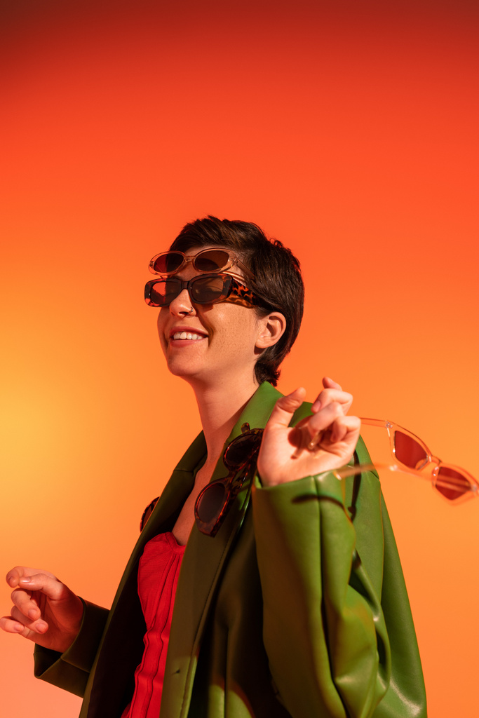 mujer joven y despreocupada posando en chaqueta verde elegante y diferentes gafas de sol de moda sobre fondo naranja - Foto, Imagen