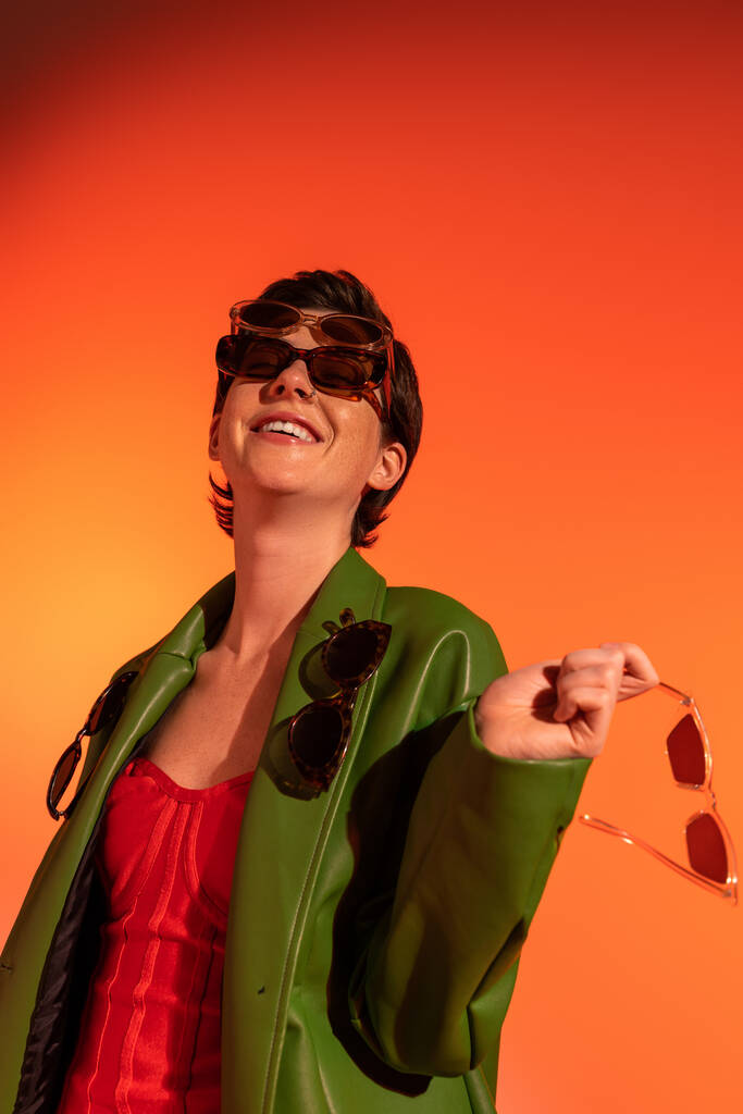 Yeşil deri ceketli, turuncu arka planda farklı güneş gözlükleriyle poz veren heyecanlı ve şık bir kadın. - Fotoğraf, Görsel