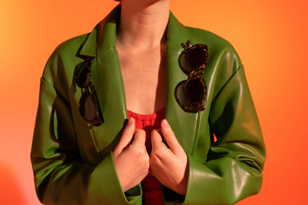 částečný pohled na ženu se stylovými slunečními brýlemi na zelené kožené bundě na oranžovém pozadí - Fotografie, Obrázek