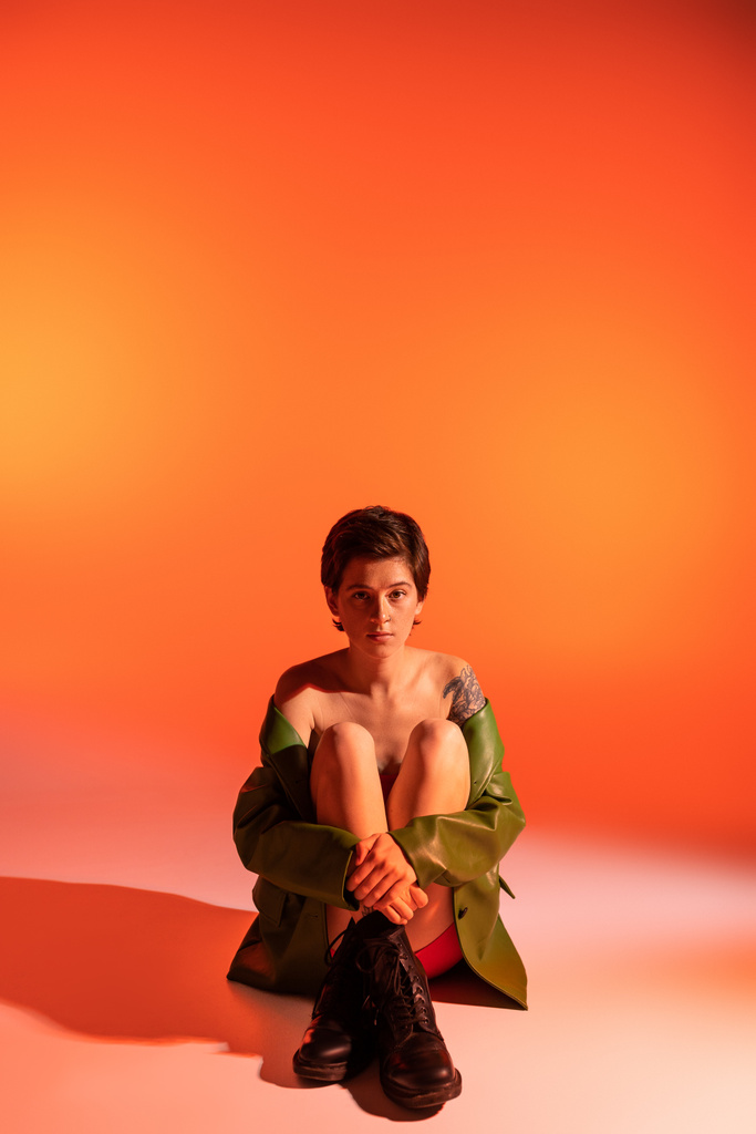 full length of brunette tattooed woman in black rough boots and green jacket sitting on orange background - Photo, Image