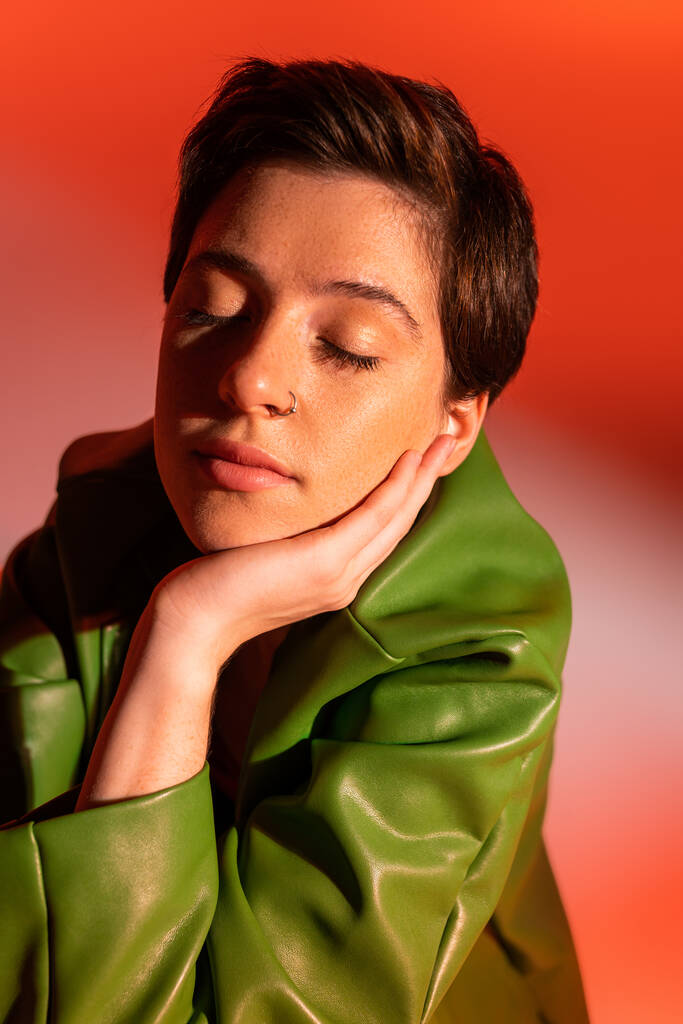 retrato de mujer morena en chaqueta de cuero verde posando con los ojos cerrados y la mano cerca de la cara sobre fondo naranja y rosa - Foto, Imagen