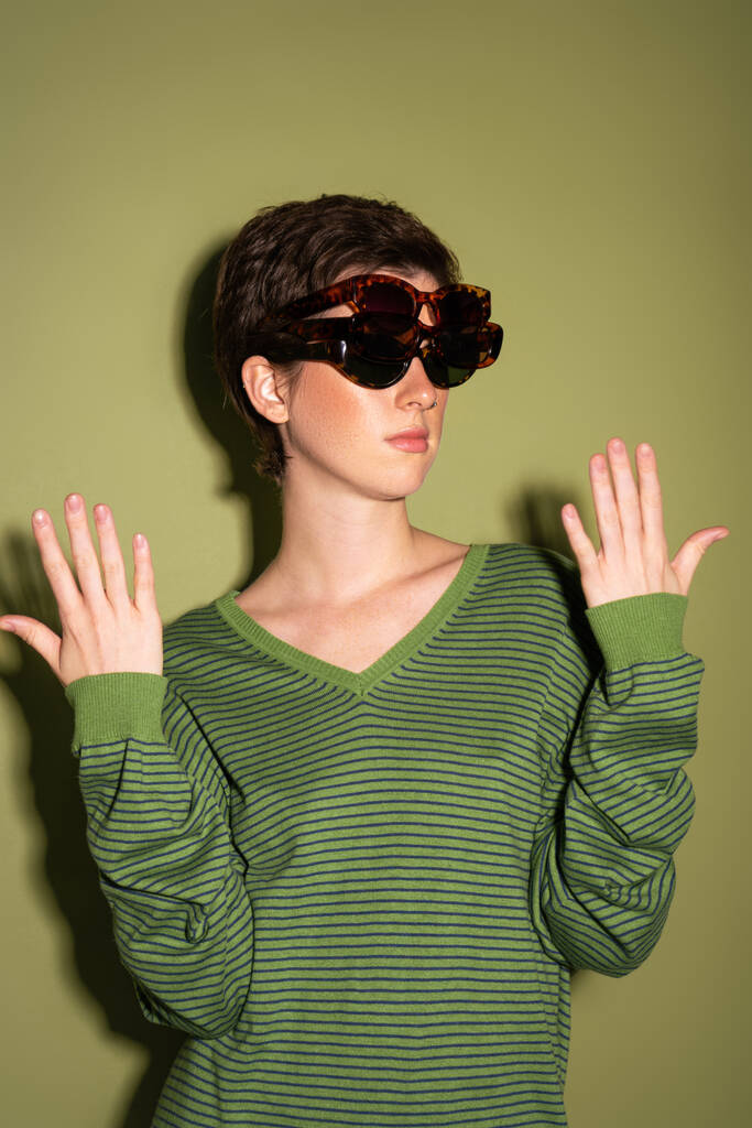 brunette woman in striped jumper wearing several trendy sunglasses on green background - Photo, Image