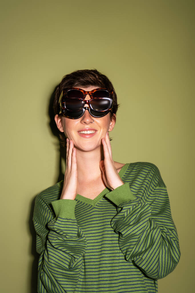 junge und fröhliche Frau posiert in gestreiftem Pullover und mehreren Sonnenbrillen auf grünem Hintergrund - Foto, Bild