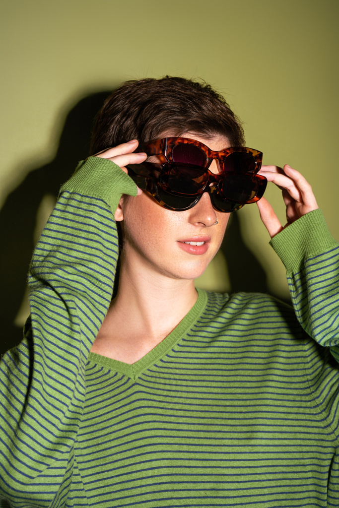 young woman in trendy striped pullover wearing several sunglasses on green background - Photo, Image