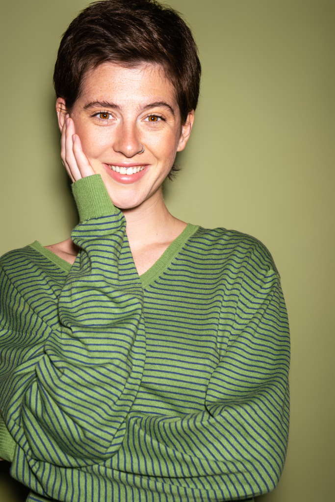 Porträt einer fröhlichen brünetten Frau in gestreiftem Pullover, die das Gesicht berührt und in die Kamera auf grünem Hintergrund blickt - Foto, Bild