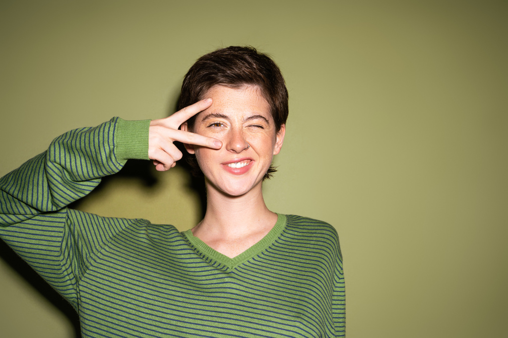 веселая женщина в полосатом свитере показывает знак победы возле лица и подмигивает в камеру на зеленом фоне - Фото, изображение
