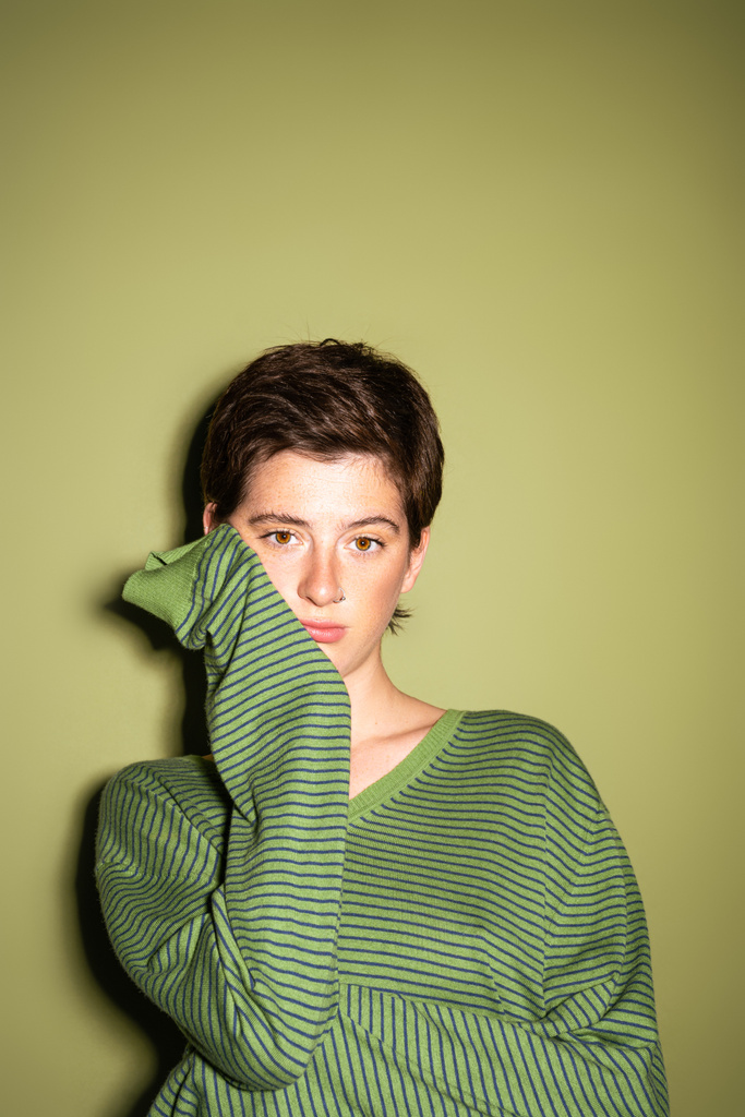 задумчивая брюнетка в модном пуловере смотрит в камеру на зеленом фоне - Фото, изображение