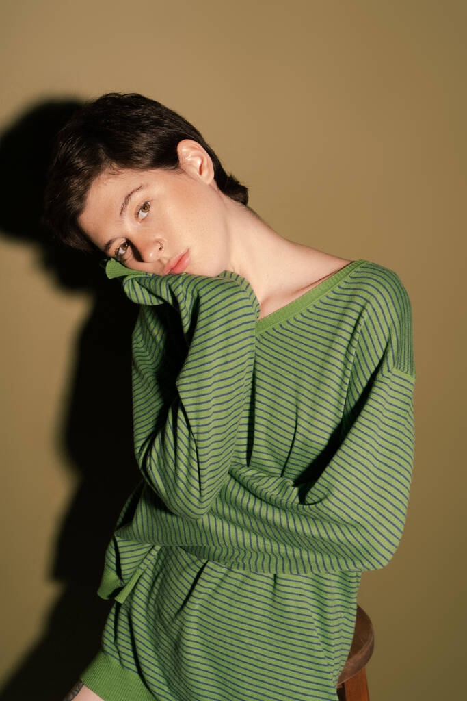 nachdenkliche Frau im gestreiften Pullover blickt in die Kamera auf grünem Hintergrund mit Schatten - Foto, Bild