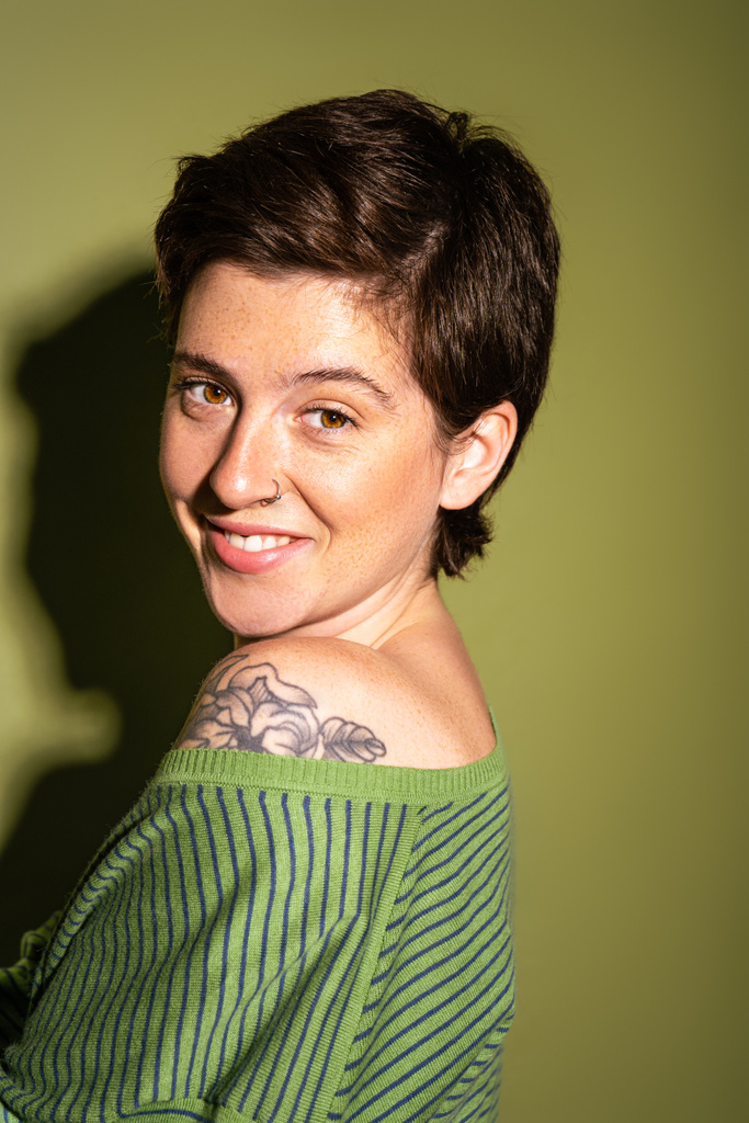 портрет радісної жінки з веснянками та татуюванням, що посміхається на камеру на зеленому фоні з тіні
 - Фото, зображення
