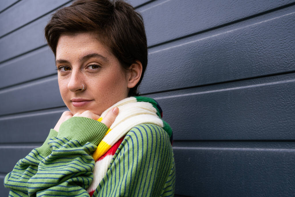 portrait of smiling freckled woman in green jumper and striped scarf standing near grey wall on street - Photo, Image