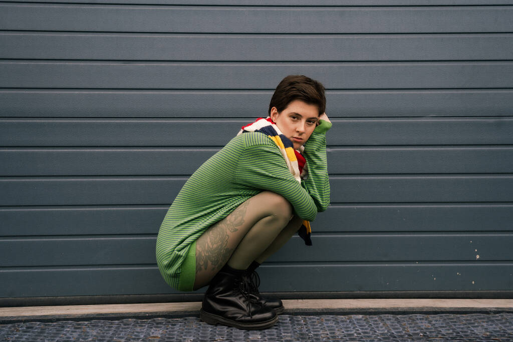 full length of trendy woman in green pullover and striped scarf sitting on haunches near grey wall on street - Photo, Image