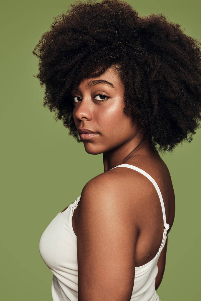 Femmina afroamericana senza emozioni in top bianco guardando la fotocamera sopra la spalla contro lo sfondo verde - Foto, immagini