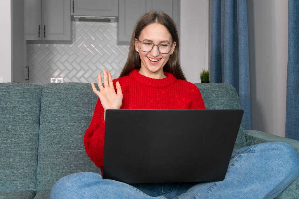 Young smiling woman in glasses, a red sweater waves her hand while sitting at a laptop on the couch. Attractive girl greets in video chat, with colleagues, friends, online communication - Photo, Image