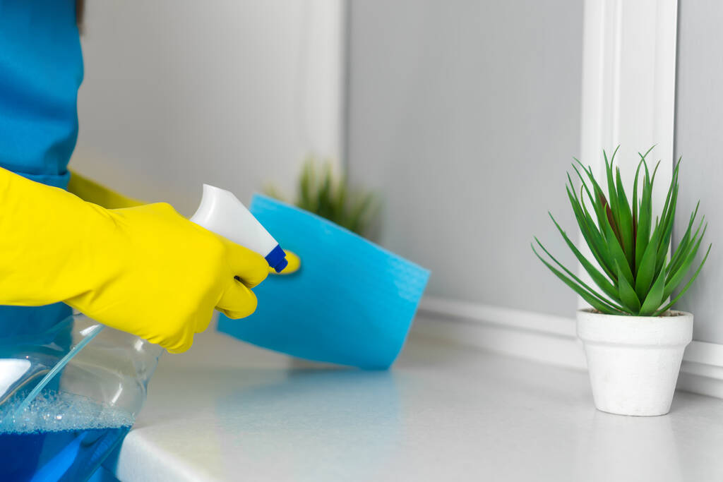 Großaufnahme weiblicher Hände in gelben Handschuhen, die ein Mikrofasertuch und Fensterputzer in der Hand halten, um eine weiße Kunststoff-Fensterbank zu wischen. Reinigung in der Wohnung durch eine Reinigungsfirma oder Hausfrau - Foto, Bild
