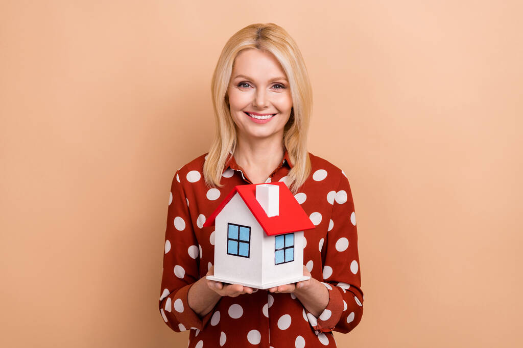 Zdjęcie przyjazne pozytywne lady zębate uśmiech ramiona trzymać pokazując mały dom odizolowany na beżowym tle koloru. - Zdjęcie, obraz