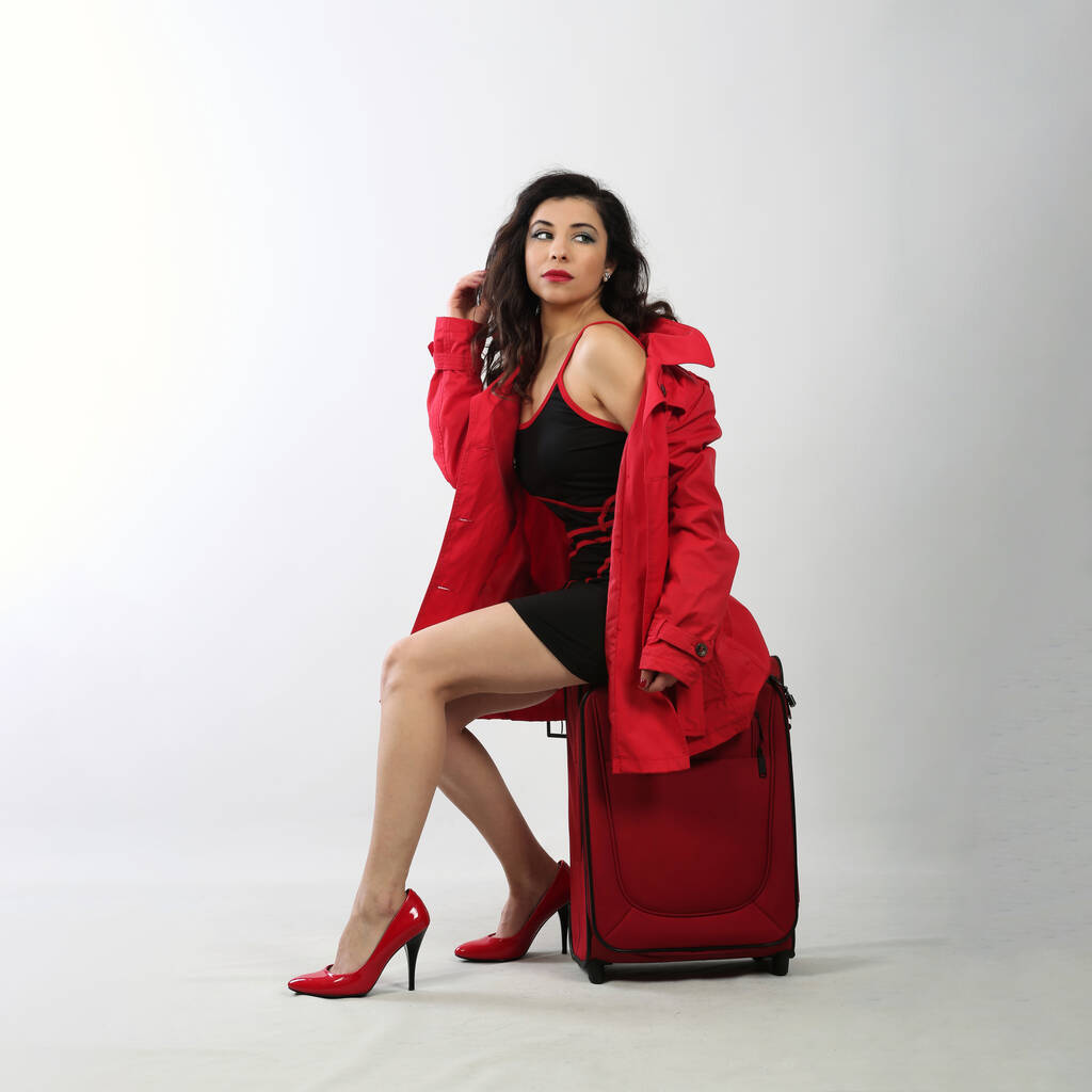 Красивая брюнетка в красном пальто с чемоданом, сидящим на чемодане - Фото, изображение