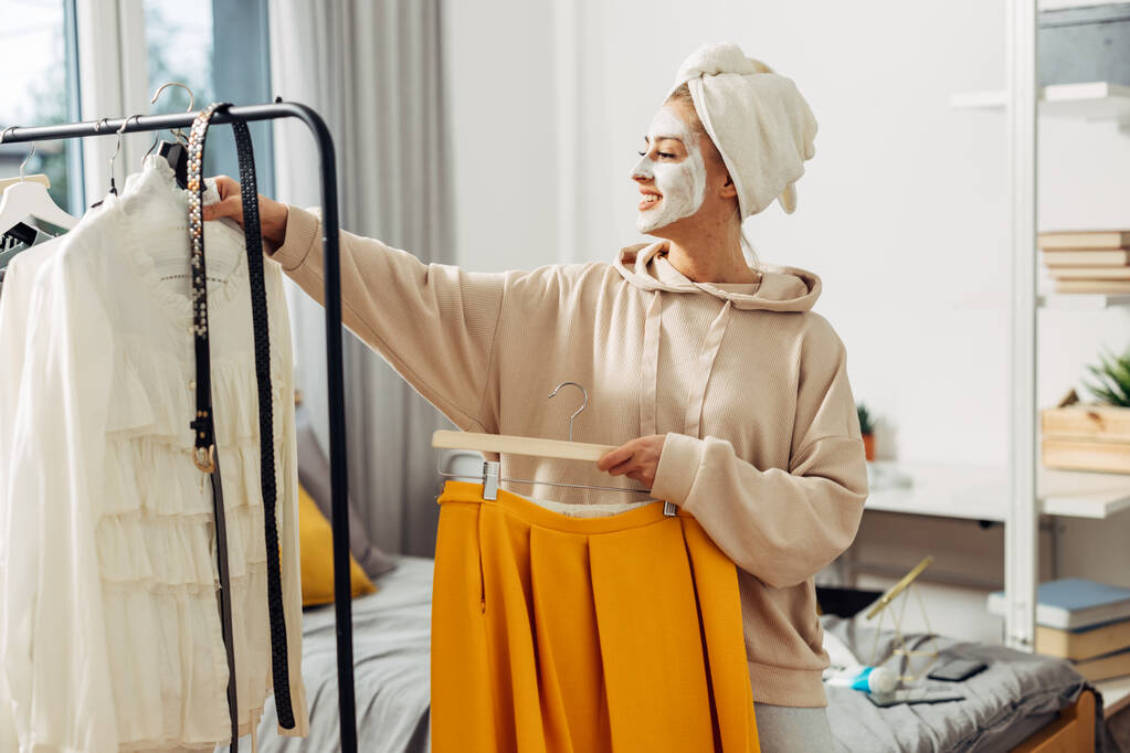 Счастливая женщина в маске и полотенце тюрбан выбирает одежду, чтобы носить - Фото, изображение