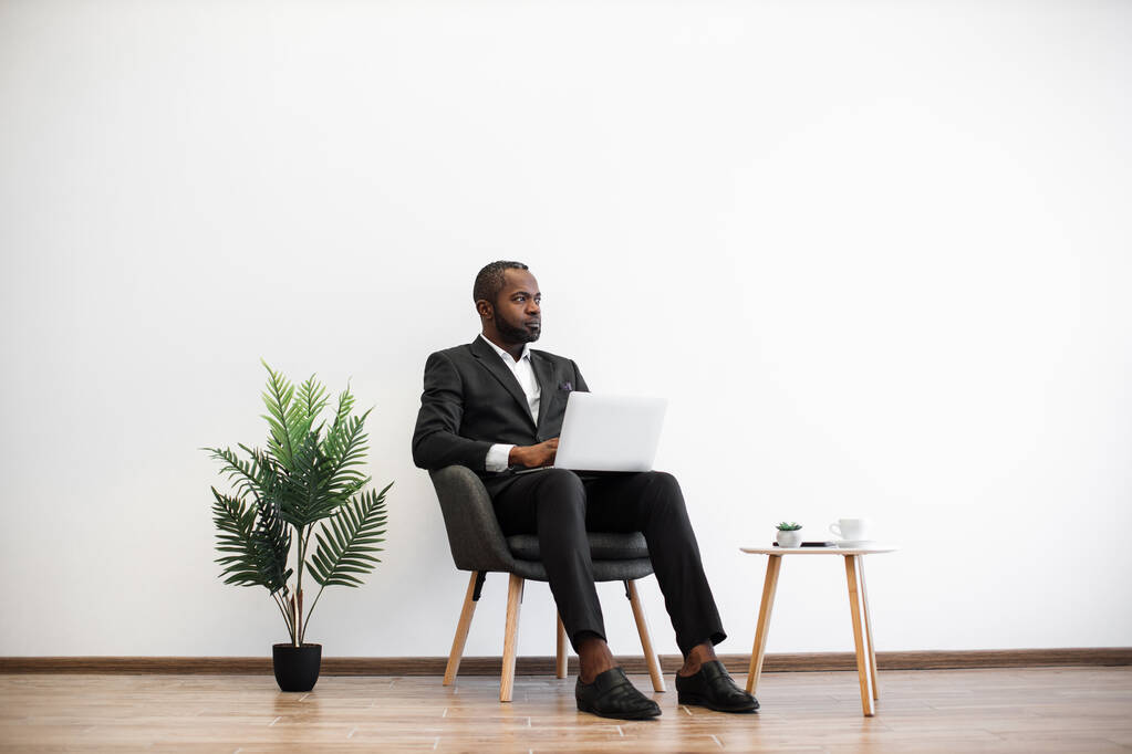 Зосереджений афроамериканець, який сидів на офісі і працював над сучасним ноутбуком. Молодий чоловік, одягнений у чорний офіційний костюм, переглядає інтернет у приміщенні. Концепція людей і технології. - Фото, зображення
