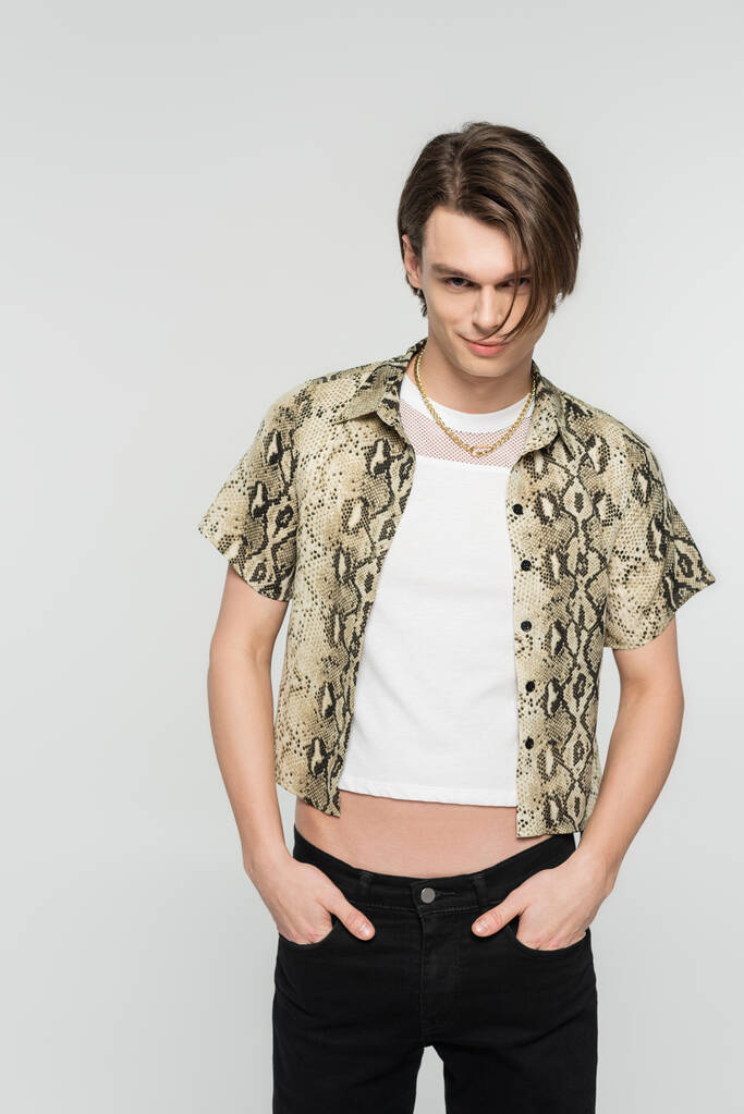 young and trendy bigender model in animal print blouse posing with hands in pockets of black pants isolated on grey - Photo, Image