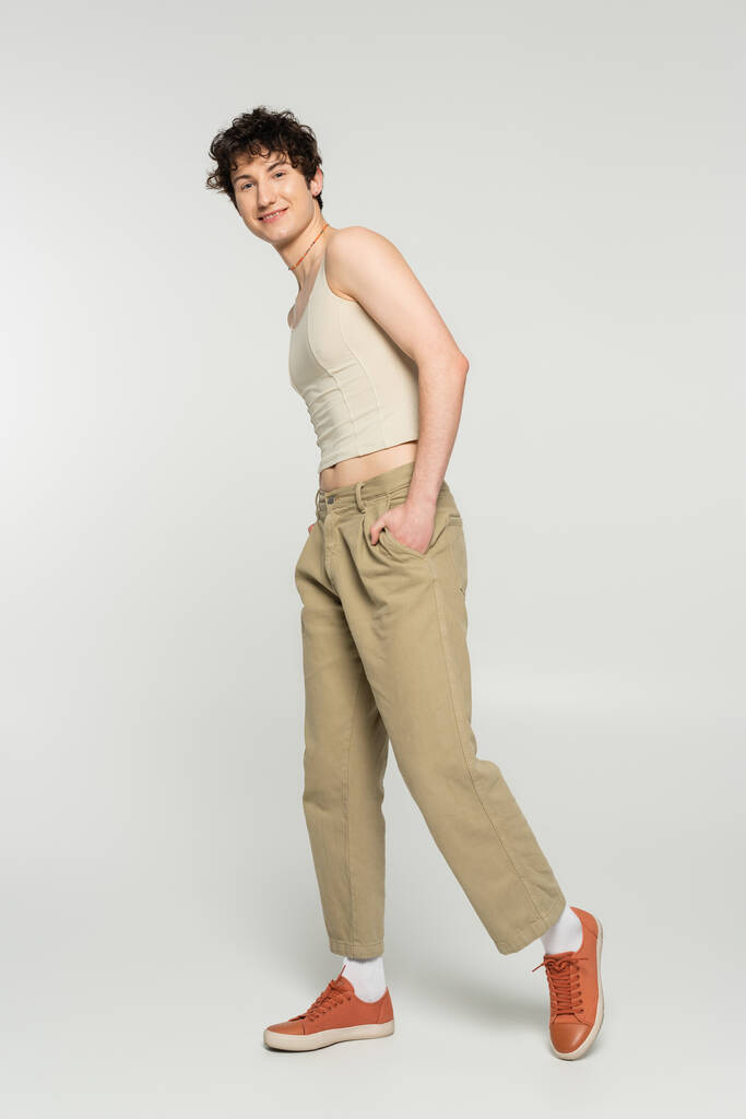 persona mayor despreocupada en camiseta sin mangas y zapatillas de deporte posando con la mano en el bolsillo de los pantalones de color beige sobre fondo gris - Foto, imagen