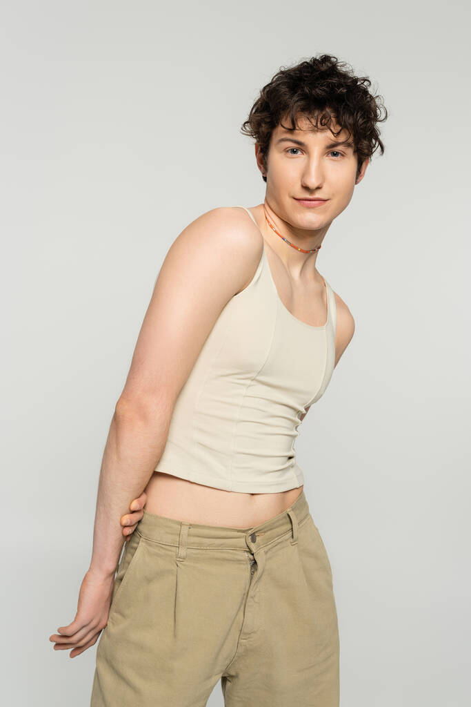 stylový pansexuální model s vlnité brunetky vlasy při pohledu na kameru izolované na šedé - Fotografie, Obrázek