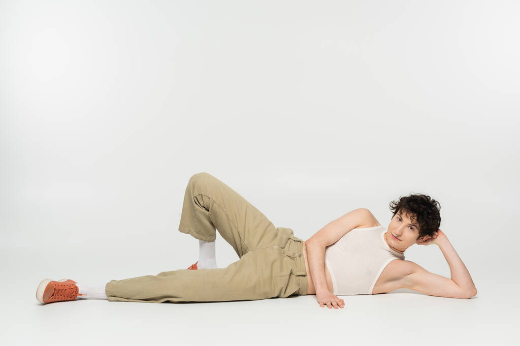 полная длина молодого пансексуала в стильной одежде лежащего и смотрящего на камеру на сером фоне - Фото, изображение