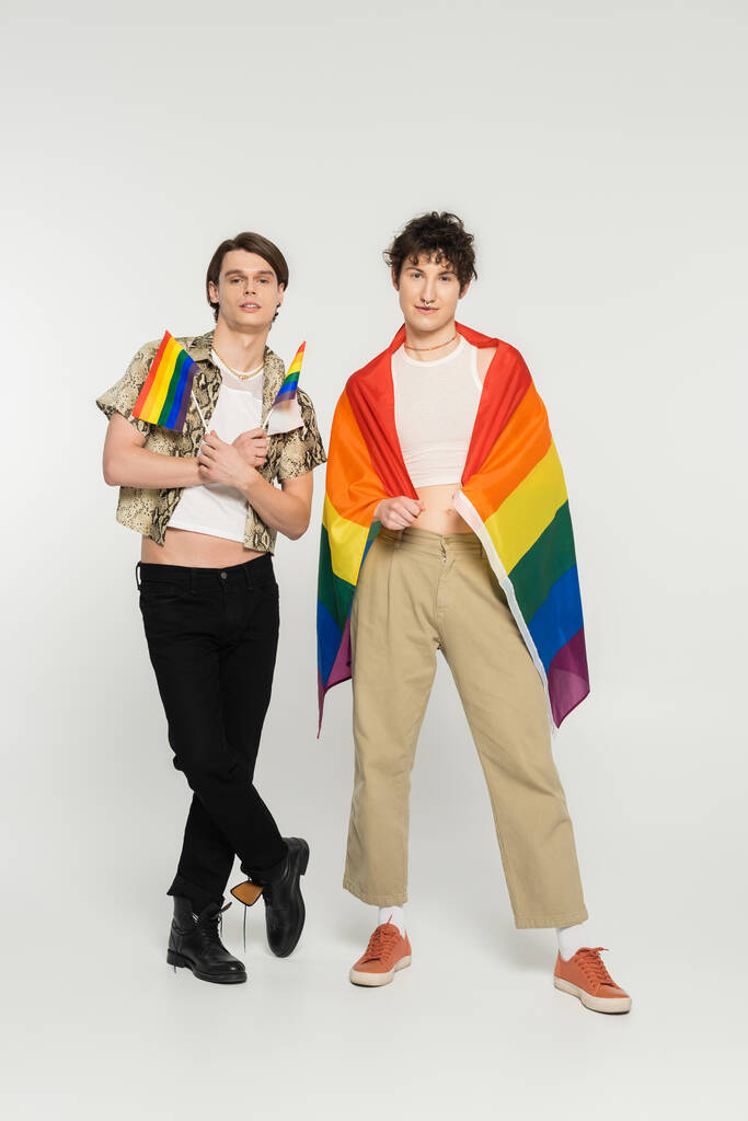 longitud completa de los jóvenes modelos no binarios en ropa elegante posando con banderas de arco iris sobre fondo gris - Foto, imagen