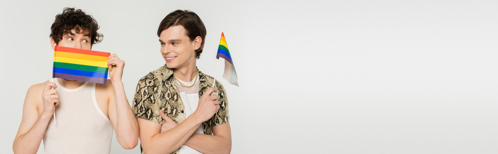χαρούμενο pansexual άτομο που κοιτάζει σύντροφο συγκαλυμμένο πρόσωπο με μικρή σημαία lgbt που απομονώνεται σε γκρι, banner - Φωτογραφία, εικόνα