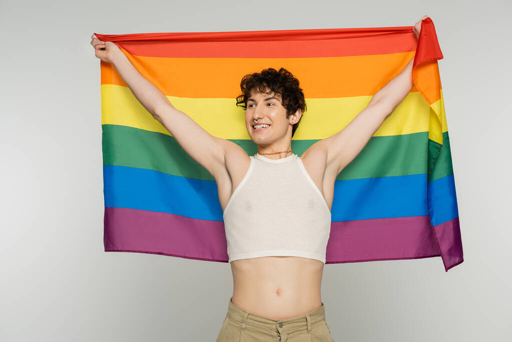 joyful bigender model in crop top holding lgbt flag in raised hands and looking away isolated on grey - Photo, Image