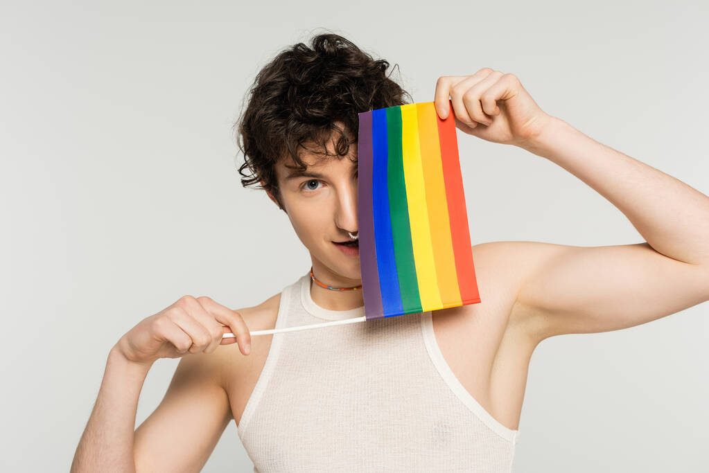 young nonbinary person obscuring face with small lgbt flag and looking at camera isolated on grey - Photo, Image