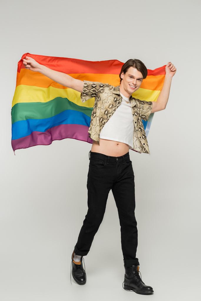 full length of stylish and carefree nonbinary person walking with rainbow flag on grey background - Photo, Image