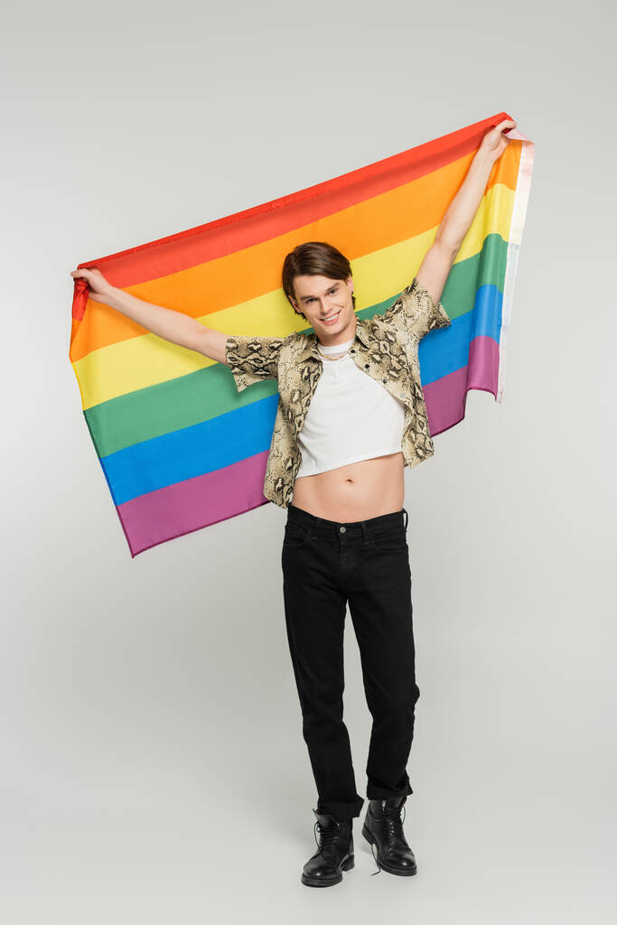 longitud completa de la persona despreocupada y de moda más grande que sostiene la bandera del arco iris en manos levantadas sobre fondo gris - Foto, Imagen