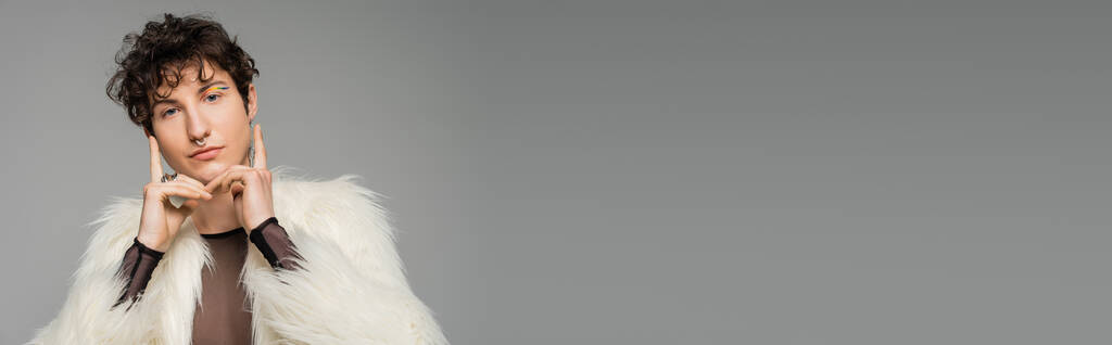 modna bigender osoba w białym faux futro kurtka trzymając się za ręce w pobliżu twarzy i patrząc na aparat odizolowany na szary, baner - Zdjęcie, obraz