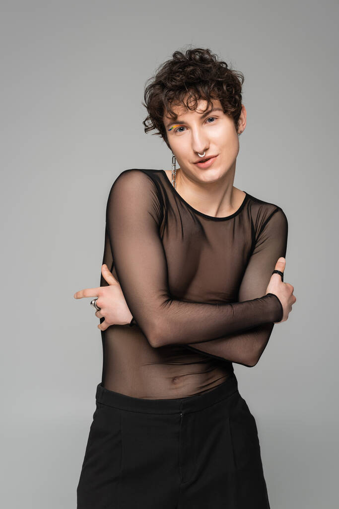 brunetka pansexual model w czarny przezroczysty top pozowanie z skrzyżowane ramiona odizolowane na szary - Zdjęcie, obraz
