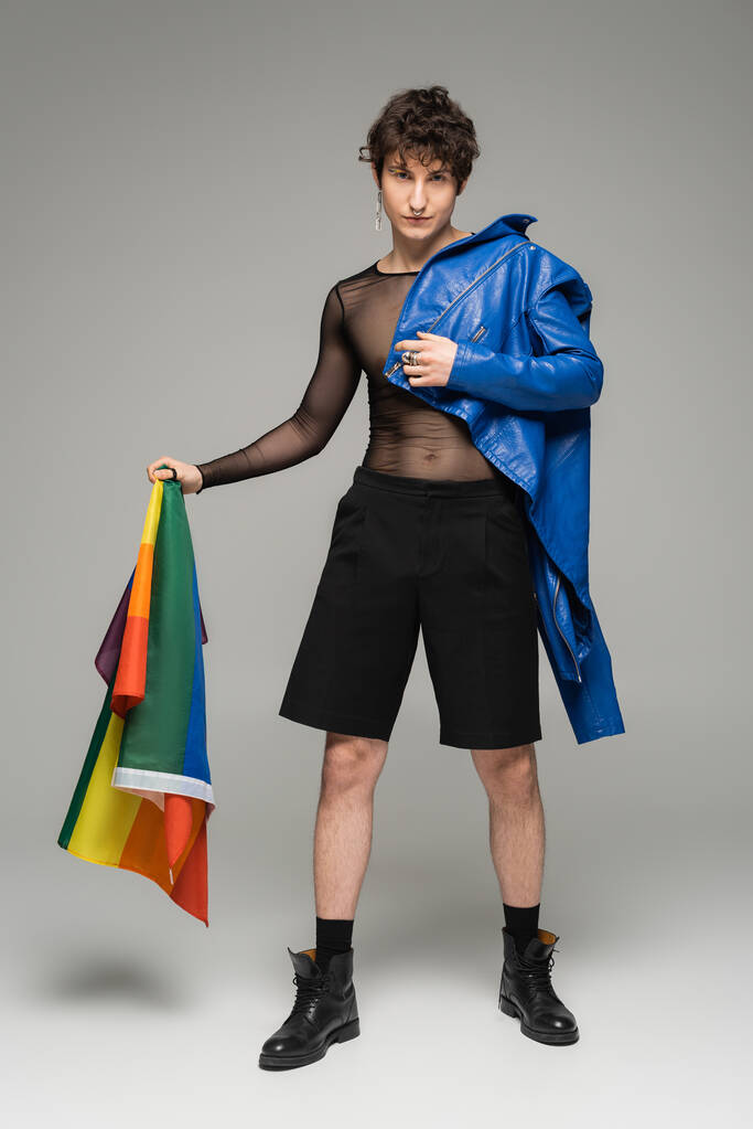 full length of trendy pangender person with blue leather jacket holding lgbt flag on grey background - Photo, Image