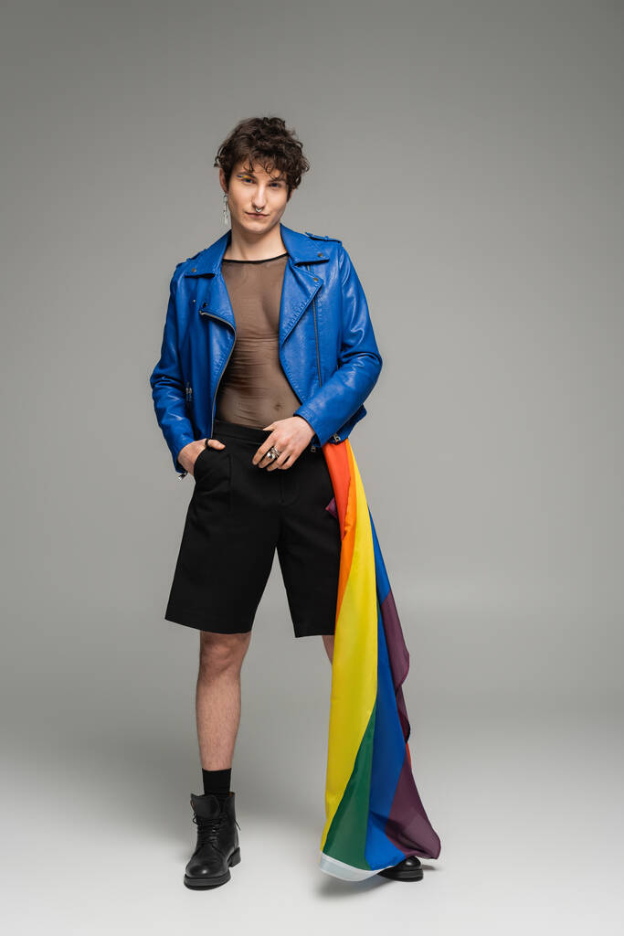 full length of pansexual person with lgbt flag posing in blue leather jacket and black shorts on grey background - Photo, Image