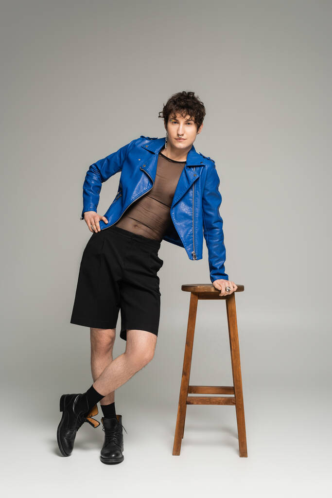 longitud completa de la persona más grande en chaqueta de cuero azul y pantalones cortos negros posando cerca de taburete de madera sobre fondo gris - Foto, Imagen