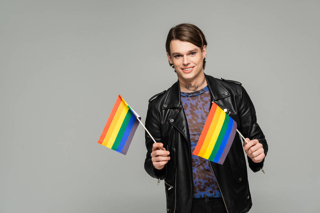 happy pangender person in black leather jacket holding small lgbt flags and smiling at camera isolated on grey - Photo, Image